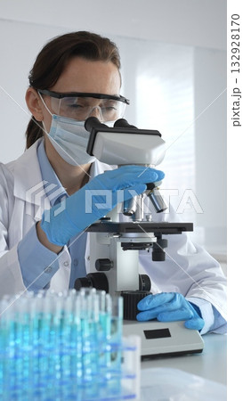 Female scientist examining microscope slide, wearing protective gear in sterile laboratory, researching pharmaceutical breakthrough with blue tinted test tube specimens Female scientist examining microscope slide, wearing protective gear in sterile laboratory, researching pharmaceutical breakthrough with blue tinted test tube specimens 132928170