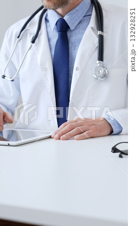Adult male doctor wearing lab coat and stethoscope using digital tablet while sitting at desk in medical office, with medical chart and pen in the foreground, close up. Medicine 132928251