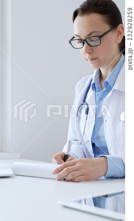 Adult doctor woman completing healthcare paperwork, clipboard positioned near laptop and stethoscope in well lit clinical workspace by window. Medicine and health care Adult doctor woman completing healthcare paperwork, clipboard positioned near laptop and stethoscope in well lit clinical workspace by window. Medicine and health care 132928259