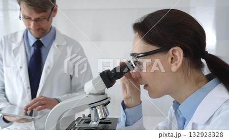 A female scientist wearing protective glasses looks into a microscope and conducts a medical research in a sterile laboratory while her male colleague works in the background. Medicine and science A female scientist wearing protective glasses looks into a microscope and conducts a medical research in a sterile laboratory while her male colleague works in the background. Medicine and science 132928328