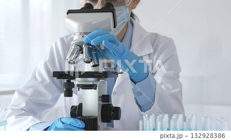 A female scientist in protective gear adjusts a microscope lens and accurately analyzes medical research samples in a sterile laboratory setting A female scientist in protective gear adjusts a microscope lens and accurately analyzes medical research samples in a sterile laboratory setting 132928386