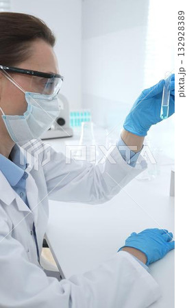 Female researcher examining clear liquid in test tube, sitting near microscopes and lab equipment within sterile scientific workspace. Medicine, healthcare and science concept 132928389