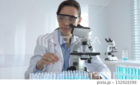 Female scientist in lab coat and safety glasses using microscope, analyzing samples in test tubes in laboratory, conducting scientific research 132928399