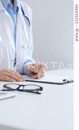 Female doctor completing healthcare paperwork, clipboard positioned near laptop and stethoscope in well lit clinical workspace by window. Medicine and health care concept Female doctor completing healthcare paperwork, clipboard positioned near laptop and stethoscope in well lit clinical workspace by window. Medicine and health care concept 132928460