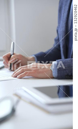 Closeup of businessman hands signing a contract with elegant pen, wearing a blue suit and a wristwatch, sitting at a white desk in office. Business people concept 132928462