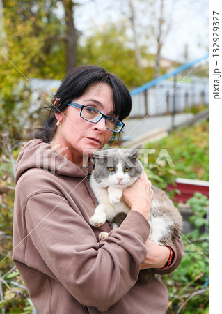 Middle aged Russian woman holding her gray and white cat outdoors in autumn garden, showing affection and calm connection with her beloved pet. Middle aged Russian woman holding her gray and white cat outdoors in autumn garden, showing affection and calm connection with her beloved pet. 132929327