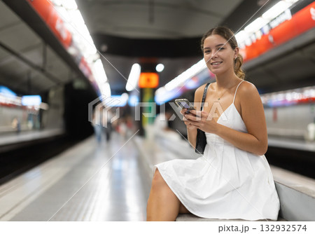 Positive young girl sitting on subway platform holding phone in hand 132932574