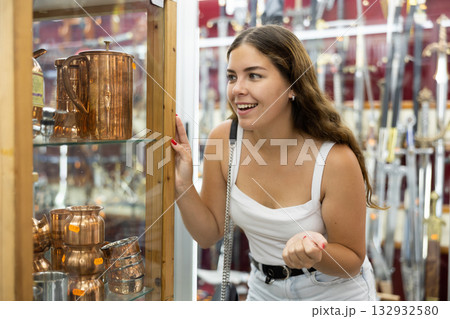 Cheerful girl looking at copper utensils in spanish souvenir shop Cheerful girl looking at copper utensils in spanish souvenir shop 132932580