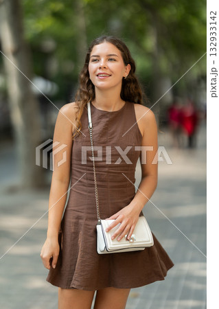 Beautiful stylish young girl in dress with white handbag walking and posing on city street on summer day Beautiful stylish young girl in dress with white handbag walking and posing on city street on summer day 132933142
