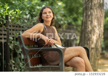 Positive cute young girl dressing in short dress sitting on bench in city park Positive cute young girl dressing in short dress sitting on bench in city park 132933226