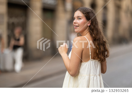 Cheerful girl in white dress walking along the street among architecture with bag 132933265