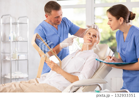 Man dermatologist examines skin of senior womans face, female nurse records data in client card 132933351