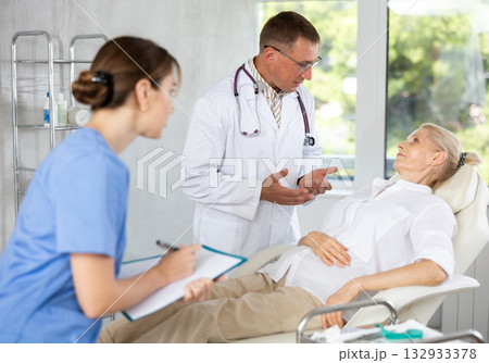 Male beautician, together with a nurse, examines the face of an mature woman and plans facelift procedures. 132933378