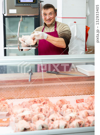 Man stands behind counter in meat department of supermarket and shows chicken carcass 132933405