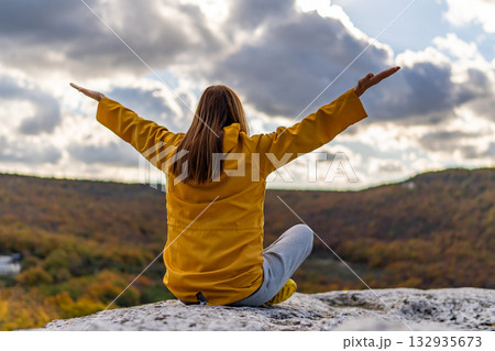 Woman nature freedom sitting on cliff rock with arms raised admiring the cloudy sky 132935673
