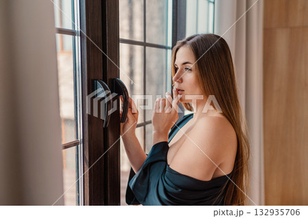 Woman, window, contemplation. Young female stands by window, touching lips in a thoughtful pose at home. Woman, window, contemplation. Young female stands by window, touching lips in a thoughtful pose at home. 132935706
