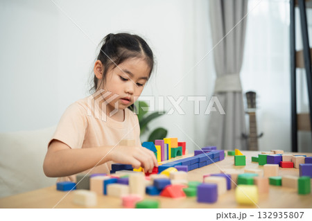 Young girl engaged in creative building play with colorful wooden blocks on a table in a bright, modern learning environment at home Young girl engaged in creative building play with colorful wooden blocks on a table in a bright, modern learning environment at home 132935807