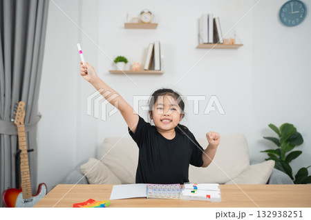 Cheerful girl in black shirt happily raising one hand holding a marker while sitting at a desk full of colorful art supplies and a cozy living room background 132938251