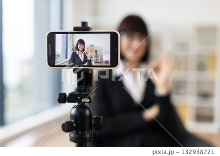 Caucasian woman wearing formal attire recording video blog using smartphone on tripod in modern office setting. Concept themes include business networking, educational content creation. Caucasian woman wearing formal attire recording video blog using smartphone on tripod in modern office setting. Concept themes include business networking, educational content creation. 132938721
