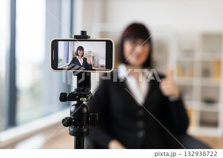 Caucasian adult woman recording herself using a smartphone on a tripod for business vlog in a bright office environment. Scene highlights communication, professionalism, and modern content creation. 132938722
