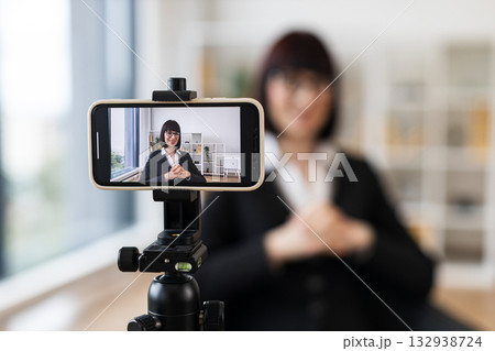 Caucasian woman in early 30s in business suit recording video content in office setting. Concept includes leadership, modern technology, and communication. Scene portrays professionalism. Caucasian woman in early 30s in business suit recording video content in office setting. Concept includes leadership, modern technology, and communication. Scene portrays professionalism. 132938724
