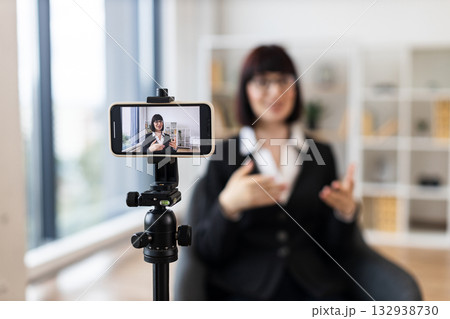 Caucasian woman recording video blog about business concepts and ideas using smartphone camera. Shows creative workspace with natural lighting for content creation and technology usage. Caucasian woman recording video blog about business concepts and ideas using smartphone camera. Shows creative workspace with natural lighting for content creation and technology usage. 132938730