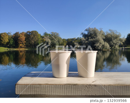 Two white paper cups are placed on a wooden table, with a view of a lake and a park. Picturesque nature. A cozy outdoor recreation. Gatchina Palace Park. Russia. 132939566