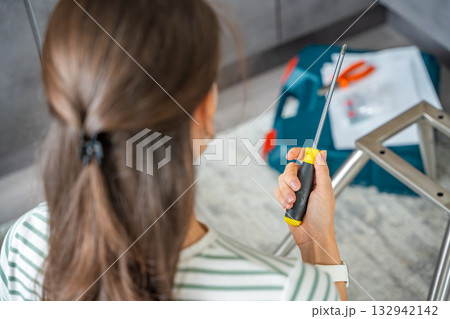 Young woman seen from behind assembling a chair with screwdrivers on the floor at home. Feminist concept of female independence and do it yourself furniture assembly. Young woman seen from behind assembling a chair with screwdrivers on the floor at home. Feminist concept of female independence and do it yourself furniture assembly. 132942142