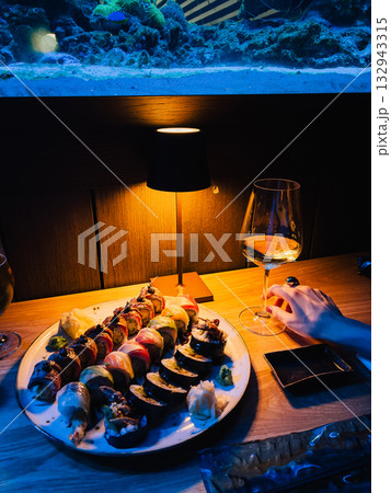 Woman hand holding glass of white wine beside plate of vibrant sushi rolls, softly illuminated by lamp in restaurant featuring large aquarium. Sophisticated dining ambiance. Glass near sushi plate Woman hand holding glass of white wine beside plate of vibrant sushi rolls, softly illuminated by lamp in restaurant featuring large aquarium. Sophisticated dining ambiance. Glass near sushi plate 132943315