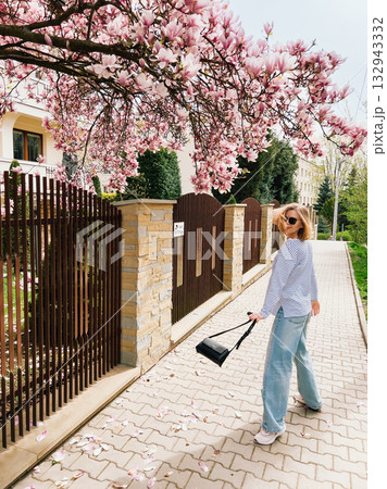 Stylish woman walking along sidewalk, surrounded by vibrant blooms of magnolia tree, savoring beauty of sunny spring day while carrying chic black purse. Blonde woman walking under magnolia blossoms Stylish woman walking along sidewalk, surrounded by vibrant blooms of magnolia tree, savoring beauty of sunny spring day while carrying chic black purse. Blonde woman walking under magnolia blossoms 132943332