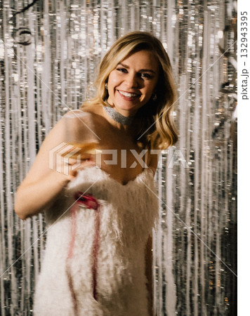 Young blond woman wearing an elegant white dress, holding glass of champagne while celebrating at New Year party, surrounded by shimmering silver curtain backdrop. Portrait of woman in New Year party 132943395