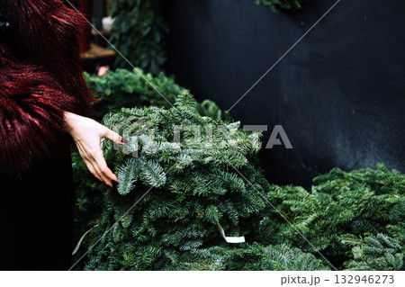 A female hand lifts a reusable pine wreath from a seasonal display. Eco-conscious decor, circular design, waste-free celebrations, nature-inspired crafting A female hand lifts a reusable pine wreath from a seasonal display. Eco-conscious decor, circular design, waste-free celebrations, nature-inspired crafting 132946273