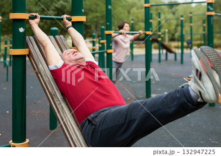 Senior man goes in for sports in city park on sports ground - doing exercises to pull his legs to chest 132947225
