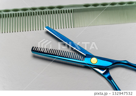 Professional hairdressing scissors with a blue handle positioned beside a green comb on a smooth surface, showcasing essential tools for hairstyling and grooming Professional hairdressing scissors with a blue handle positioned beside a green comb on a smooth surface, showcasing essential tools for hairstyling and grooming 132947532