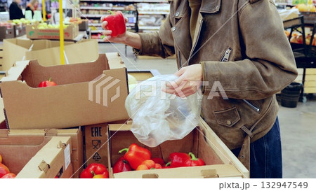 Woman in a leather jacket picks bell peppers from cardboard boxes in a grocery store, while opening a plastic bag Woman in a leather jacket picks bell peppers from cardboard boxes in a grocery store, while opening a plastic bag 132947549