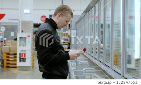 Shopper examines product labels while comparing various frozen food options in the supermarket's refrigerated aisle. A man thoughtfully chooses products in a grocery store 132947653