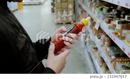 Man meticulously studies hot sauce nutritional information in store aisle, contemplating purchase surrounded by shelves of products. A customer in black studies the label on a bottle of sauce 132947657