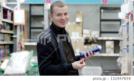 Man happily compares hair care products in a supermarket aisle, reading labels and ingredients with dedication. Background is blurred, creating cozy shopping atmosphere. 132947659