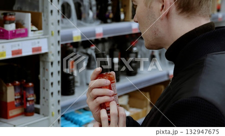 Man compares hot sauce bottles in a busy supermarket aisle, surrounded by shelves filled with products. A shopper in black studies the label on a bottle of hot sauce in the condiment section 132947675