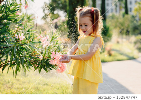 Happy little girl in yellow dress playing near sprinkler on sunny day in blooming summer park. Happy little girl in yellow dress playing near sprinkler on sunny day in blooming summer park. 132947747
