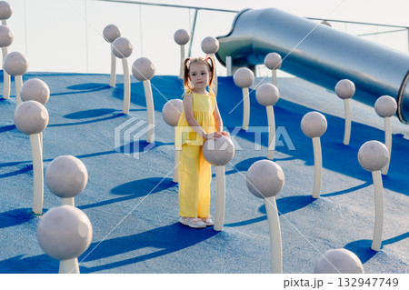 Little cute girl in yellow outfit playing on modern playground with climbing structures on a sunny day. 132947749