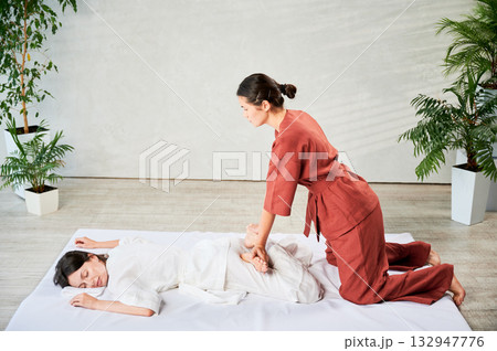 Masseuse making thai yoga massage. Woman therapist doing traditional massage treatment, stretching female patient's body. Client lying on mat, enjoying hand massage in calm atmosphere. Masseuse making thai yoga massage. Woman therapist doing traditional massage treatment, stretching female patient's body. Client lying on mat, enjoying hand massage in calm atmosphere. 132947776