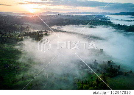 Aerial view of sunrise over mist-covered landscape, with golden light illuminating rolling hills and valleys. Thick fog creates mystical atmosphere, partially obscuring lush greenery and trees below. 132947839