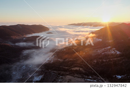 Setting sun casts golden hue over vast sea of clouds, blanketing valleys between forested hills and distant mountain ranges. Aerial view of tranquil scene captures serene beauty of nature. Setting sun casts golden hue over vast sea of clouds, blanketing valleys between forested hills and distant mountain ranges. Aerial view of tranquil scene captures serene beauty of nature. 132947842