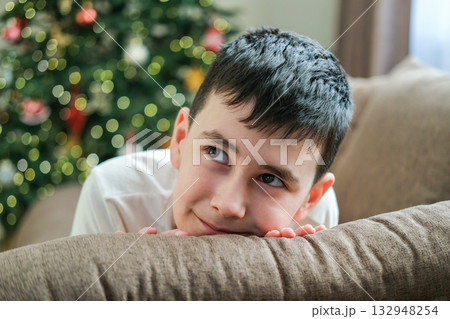 Portrait of cute nine-year-old boy looking up dreamily and smiling slightly against backdrop of Christmas tree and glowing lights in anticipation of holiday and gifts Portrait of cute nine-year-old boy looking up dreamily and smiling slightly against backdrop of Christmas tree and glowing lights in anticipation of holiday and gifts 132948254