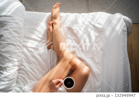 Close-up of female legs with cup of coffee on bed. Close-up of female legs with cup of coffee on bed. 132949947