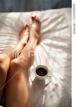 Close-up of female legs with cup of coffee on bed. Close-up of female legs with cup of coffee on bed. 132949949