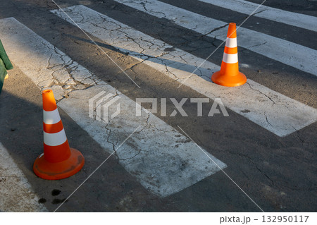 Two orange traffic cones are placed next to a pedestrian crosswalk on a cracked pavement as shadows stretch in the afternoon light 132950117