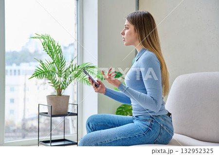 Young woman sitting at home in chair using smartphone, having video chat Young woman sitting at home in chair using smartphone, having video chat 132952325