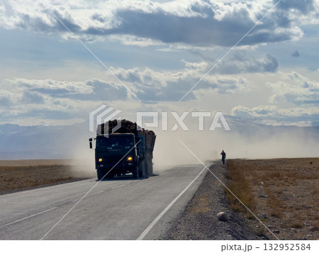 Log truck on dusty rural highway kicks up dust with person walking along road open landscape distant mountains dramatic clouds Log truck on dusty rural highway kicks up dust with person walking along road open landscape distant mountains dramatic clouds 132952584